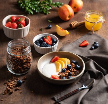 Sağlıklı ve besleyici kahvaltı kavramı. Ev yapımı granola, şeftali, çilek, bal ve yabanmersinli lezzetli yoğurt, koyu ahşap bir arka planda. Diyet yemek..