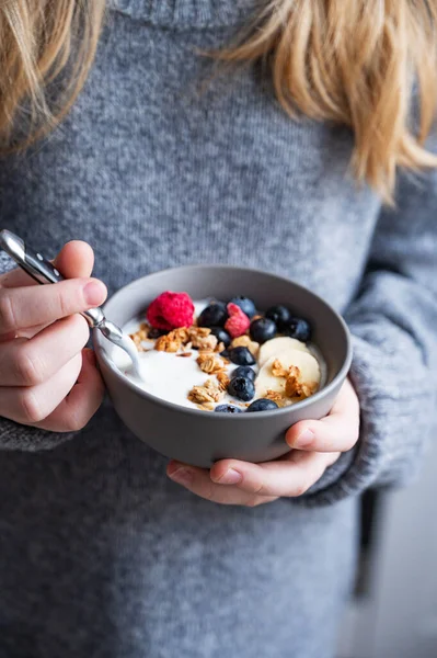 a young girl in a gray sweater is holding a bowl of vegetarian yogurt, granola and fresh blueberry and banana. The concept of a healthy breakfast.
