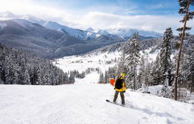 Arkhyz kayak merkezinin güzel manzarası. Dağları, karı, ormanı ve güneşli bir kış gününde, sarı ceketli ve sırt çantalı bir snowboardcu. Kafkas Dağları, Rusya.