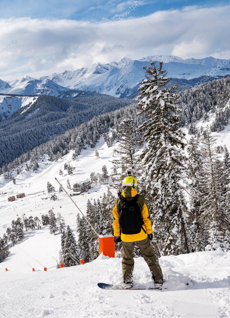 Beautiful landscape of the Arkhyz ski resort with mountains, snow, forest and man snowboarder in yellow jacket and backpack on a sunny winter day.  Caucasus  Mountains, Russia.