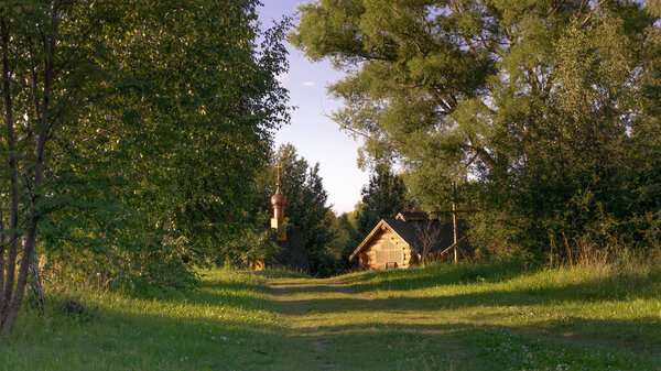 A forest road leads to a wooden structure and an Orthodox chapel. There are large deciduous trees near the road.