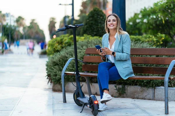 Stylish businesswoman using smartphone on bench near electric scooter — Stock Photo, Image