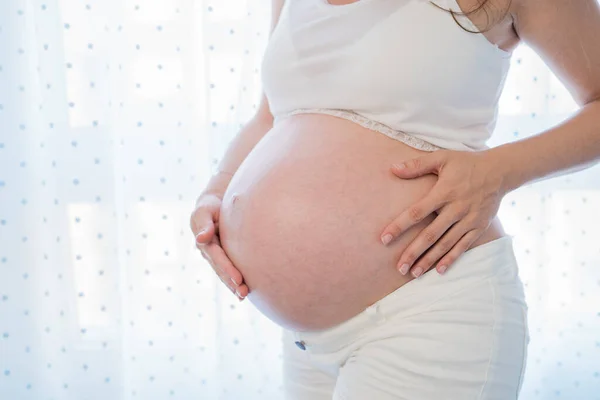 Ansiktslös gravid kvinna står nära fönstret och vidrör magen — Stockfoto