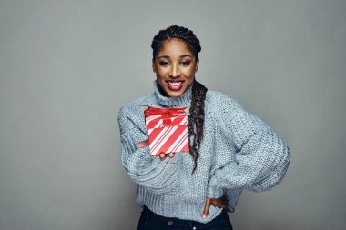 Gri arka planda kırmızı çizgili Noel hediyesi veren eli kalçasında gülümseyen genç bir kadının portresi.