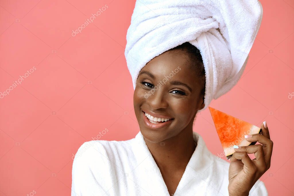 Young beautiful woman in bathrobe with white towel on head holding slice of watermelon on pink background