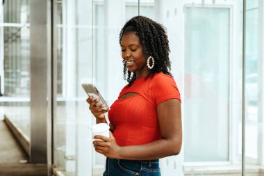 Genç Afro-Amerikan kadın akıllı telefon kullanıyor, tek kullanımlık bardak tutuyor ve gülümsüyor.