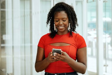 Kırmızı bluzlu mutlu Afro-Amerikan kadın akıllı telefon kullanıyor.