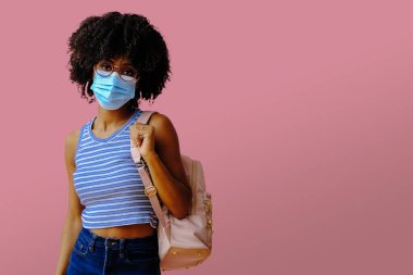 Sırt çantalı, koruyucu maskeli ve pembe arka planda poz veren genç kız öğrenci okula geri dönüyor. 