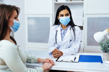 Maskeli doktor ve hasta sağlık merkezindeki masada otururken sağlık sorunlarını tartışıyorlar. 