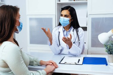 Maskeli doktor ve hasta sağlık merkezindeki masada otururken sağlık sorunlarını tartışıyorlar. 