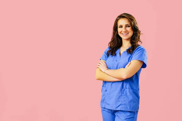 Gülümseyen kadın doktor portresi ya da pembe stüdyo arka planında kollarını kavuşturmuş mavi üniformalı hemşire.