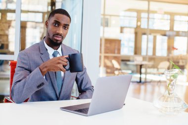 Genç bir Afrikalı Amerikalı iş adamının portresi masada dizüstü bilgisayarla oturuyor ve kahve içiyor.