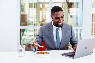 Afro-Amerikan iş adamı ofiste dizüstü bilgisayarla çalışırken yemek yiyor. 