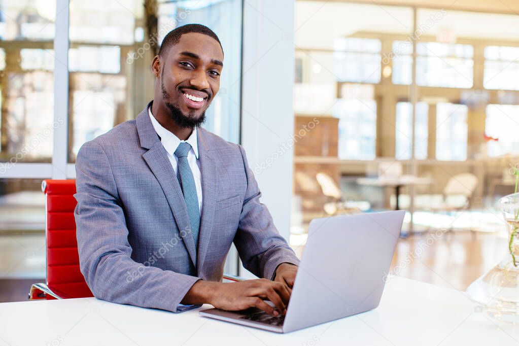 portrait of a smiling african american businessman using laptop sitting at the table and looking at camera