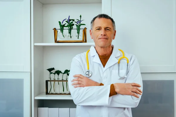 Erkek doktor steteskopla ayakta duruyor ve kollarını kavuşturup gülümsüyor.