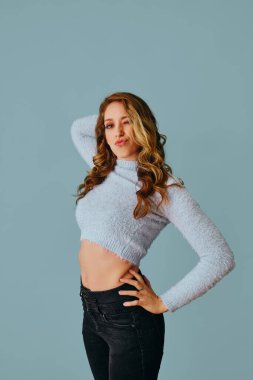 Gri arka planda kazak ve kot pantolon giyen güzel genç bir kadın.