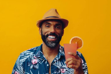 Havai gömleği ve sarı arka planda portakal suyu pozu veren şapkalı yakışıklı Afro-Amerikan bir adam.