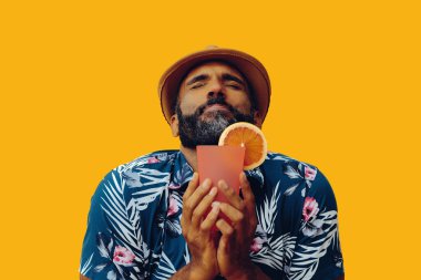 Havai gömleği ve sarı arka planda portakal suyu pozu veren şapkalı yakışıklı Afro-Amerikan bir adam.