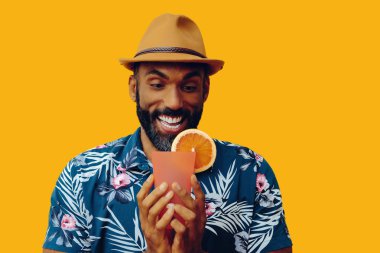 Havai gömleği ve sarı arka planda portakal suyu pozu veren şapkalı yakışıklı Afro-Amerikan bir adam.