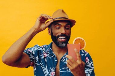 Havai gömleği ve sarı arka planda portakal suyu pozu veren şapkalı yakışıklı Afro-Amerikan bir adam.