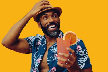 Havai gömleği ve sarı arka planda portakal suyu pozu veren şapkalı yakışıklı Afro-Amerikan bir adam.