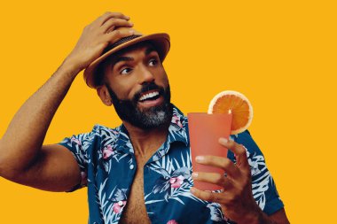 Havai gömleği ve sarı arka planda portakal suyu pozu veren şapkalı yakışıklı Afro-Amerikan bir adam.