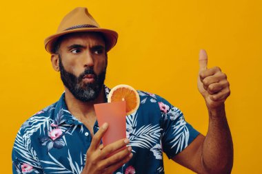 Havai gömleği ve sarı arka planda portakal suyu pozu veren şapkalı yakışıklı Afro-Amerikan bir adam.