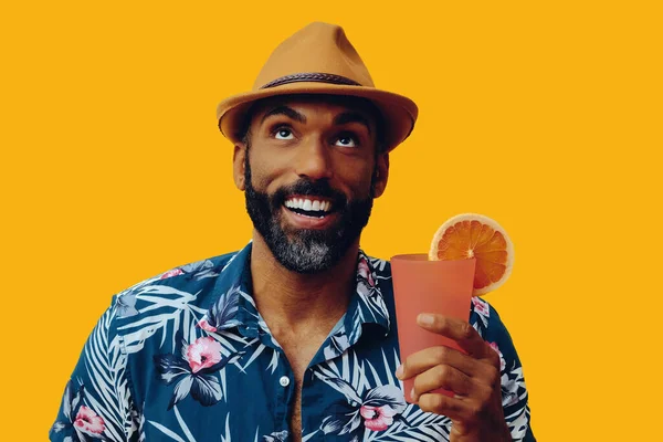Havai gömleği ve sarı arka planda portakal suyu pozu veren şapkalı yakışıklı Afro-Amerikan bir adam.