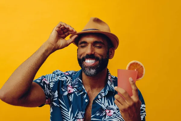 Havai gömleği ve sarı arka planda portakal suyu pozu veren şapkalı yakışıklı Afro-Amerikan bir adam.
