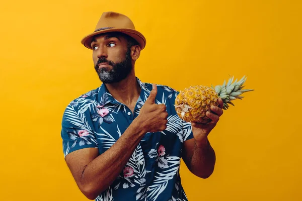 Hawaii gömlekli yakışıklı Afro-Amerikalı adam sarı arka planda ananasla poz veriyor.