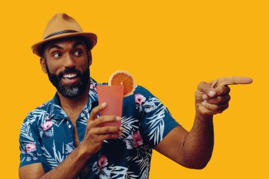 Havai gömleği ve sarı arka planda portakal suyu pozu veren şapkalı yakışıklı Afro-Amerikan bir adam.