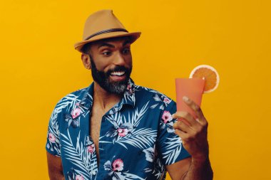 Havai gömleği ve sarı arka planda portakal suyu pozu veren şapkalı yakışıklı Afro-Amerikan bir adam.