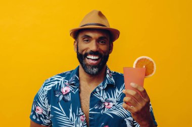 Havai gömleği ve sarı arka planda portakal suyu pozu veren şapkalı yakışıklı Afro-Amerikan bir adam.