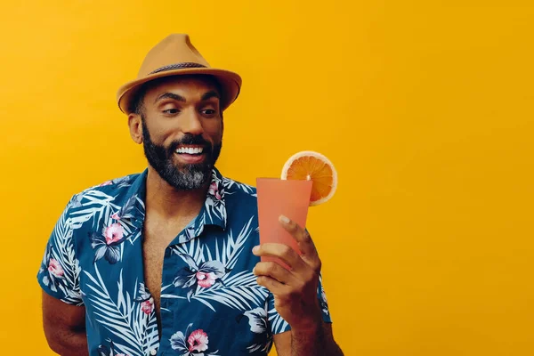 Havai gömleği ve sarı arka planda portakal suyu pozu veren şapkalı yakışıklı Afro-Amerikan bir adam.