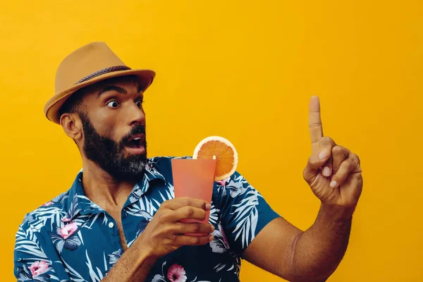 Havai gömleği ve sarı arka planda portakal suyu pozu veren şapkalı yakışıklı Afro-Amerikan bir adam.