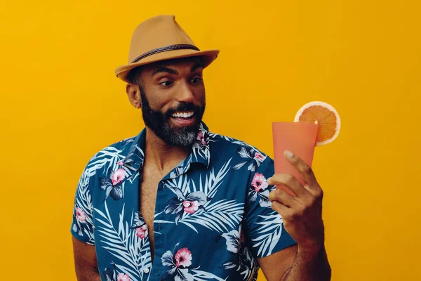 Havai gömleği ve sarı arka planda portakal suyu pozu veren şapkalı yakışıklı Afro-Amerikan bir adam.