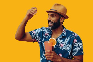 Havai gömleği ve sarı arka planda portakal suyu pozu veren şapkalı yakışıklı Afro-Amerikan bir adam.