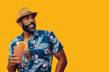 Havai gömleği ve sarı arka planda portakal suyu pozu veren şapkalı yakışıklı Afro-Amerikan bir adam.