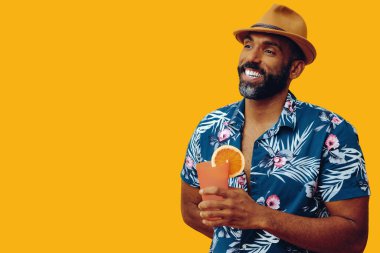 Havai gömleği ve sarı arka planda portakal suyu pozu veren şapkalı yakışıklı Afro-Amerikan bir adam.
