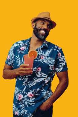 Havai gömleği ve sarı arka planda portakal suyu pozu veren şapkalı yakışıklı Afro-Amerikan bir adam.