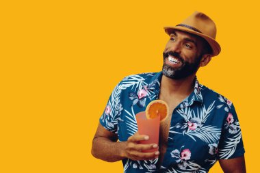 Havai gömleği ve sarı arka planda portakal suyu pozu veren şapkalı yakışıklı Afro-Amerikan bir adam.