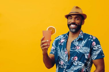 Havai gömleği ve sarı arka planda portakal suyu pozu veren şapkalı yakışıklı Afro-Amerikan bir adam.