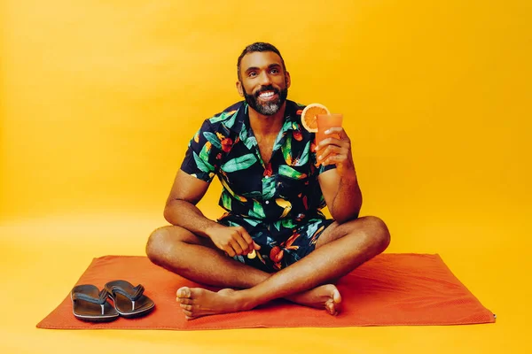 handsome african american man on vacation sitting on an orange towel, holding orange juice