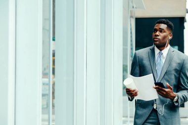 Young adult entrepreneur using smartphone outside wearing suit, tie and headphones holding documents