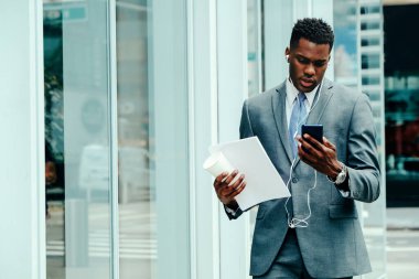 Young adult businessman using smartphone outside wearing suit, tie and headphones holding documents