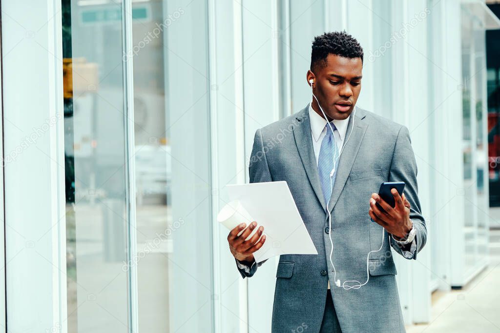 Young adult entrepreneur using smartphone outside wearing suit, tie and headphones holding documents