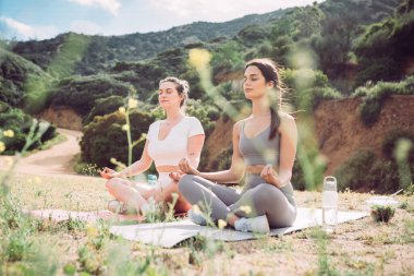 Two women in workout clothing meditating in rugged, outdoor setting