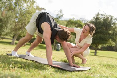 Two millennial friends practicing yoga in the park, one correcting the other's form
