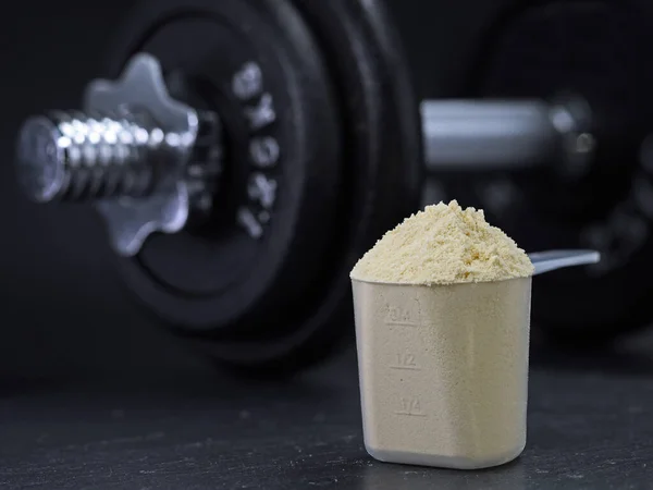 Measuring scoop filled with whey protein powder with dumbbell on black background, protein shake muscle building.