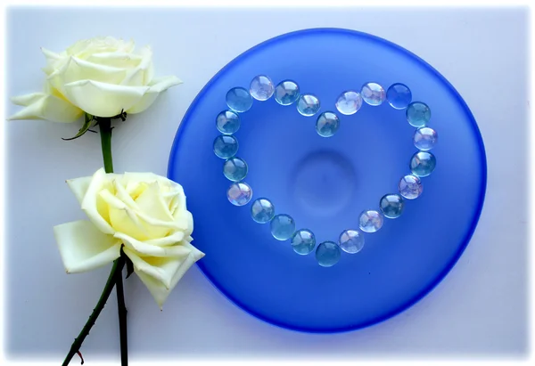 Deux roses blanches avec un cœur de perles de verre sur plaque de verre bleu — Photo